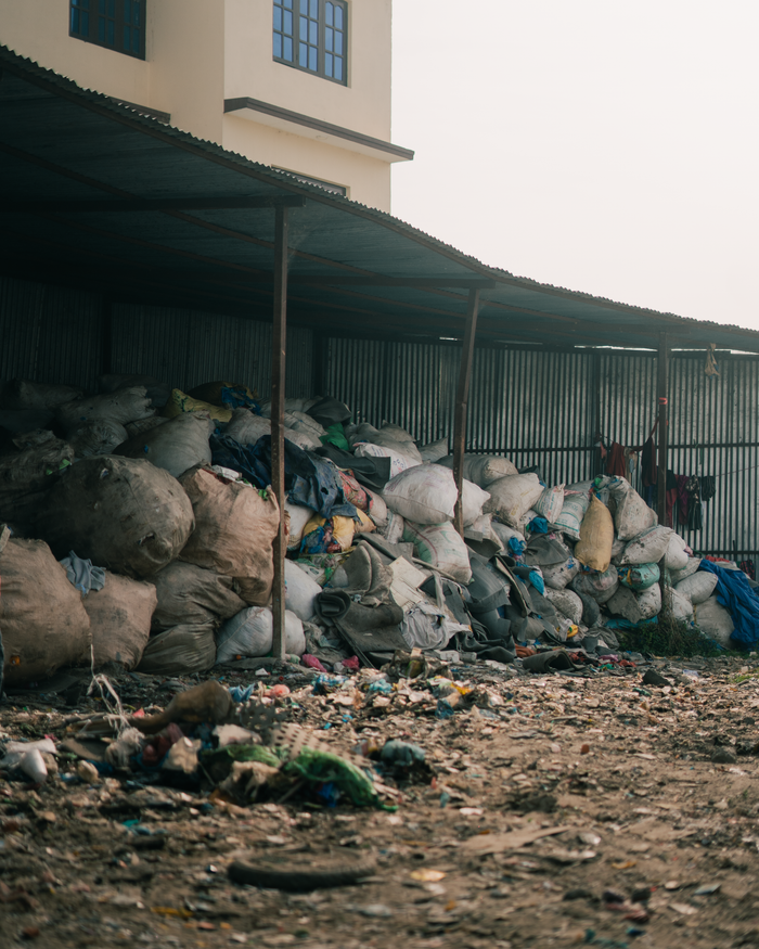 About Piles of garbage bags stacked in an outdoor waste disposal area near industrial buildings.