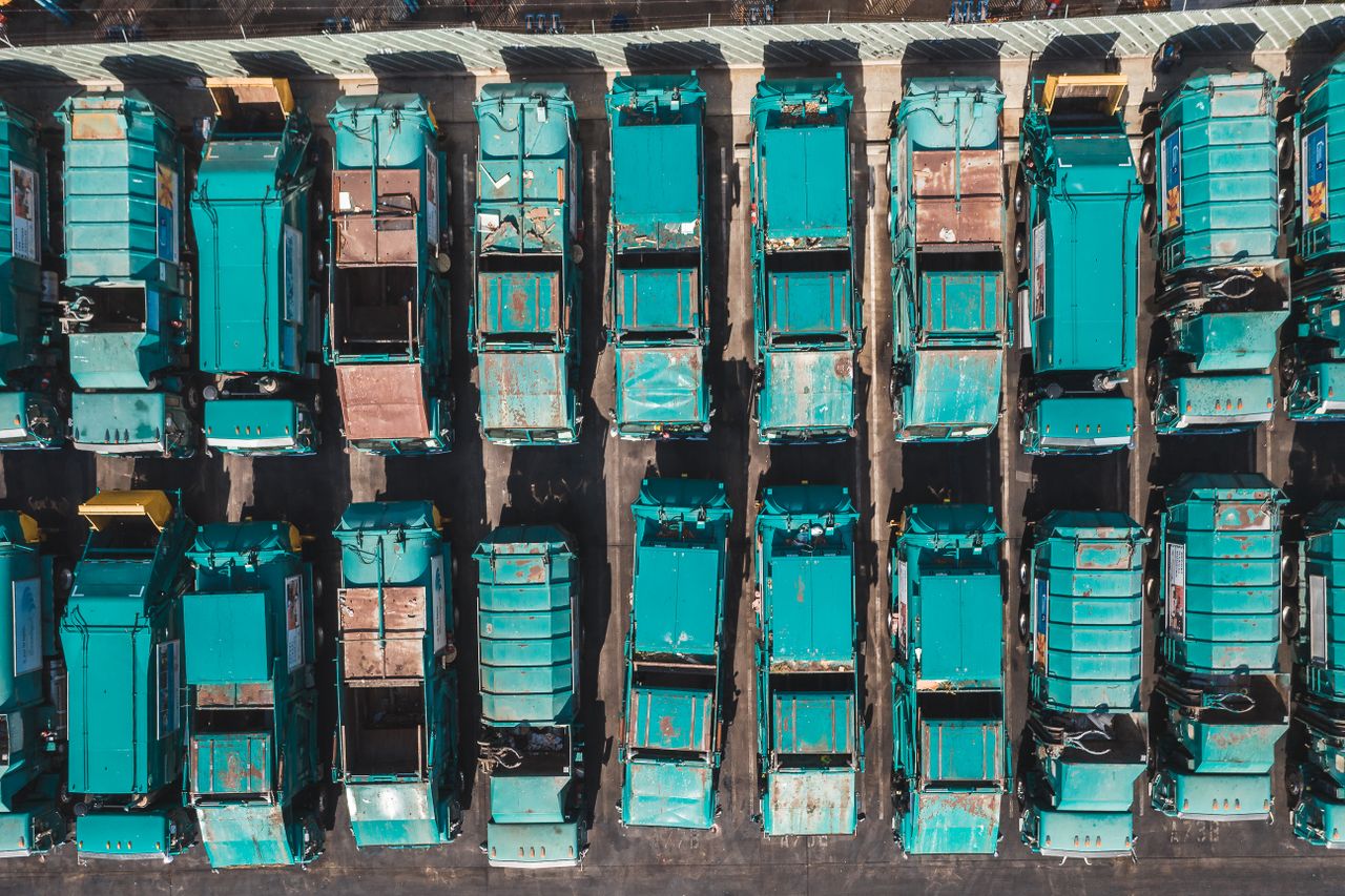 Services Aerial shot of turquoise garbage trucks parked in neat rows, showcasing a pattern of urban utility vehicles.